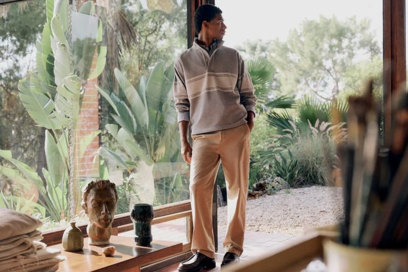 A dark-skinned man stands indoors near a window wearing a grey, cream, and brown striped collared shirt and tan trousers in a fashion editorial.