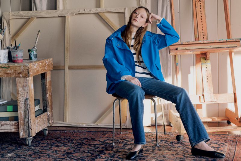 A woman wearing a blue jacket and jeans sits on a stool in an artist's studio, posing near a canvas and painting supplies.