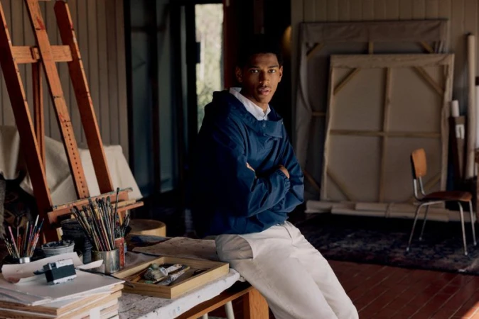 A man in a blue jacket leaning against an art studio table with paintbrushes and easels in the background