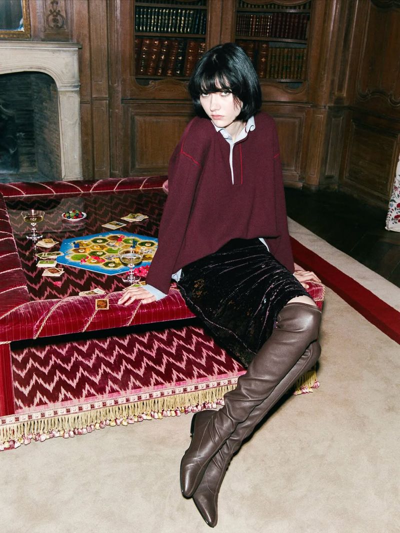 A brunette woman poses indoors, wearing a burgundy top, black skirt, and brown boots, seated on a red patterned couch in front of a bookcase.