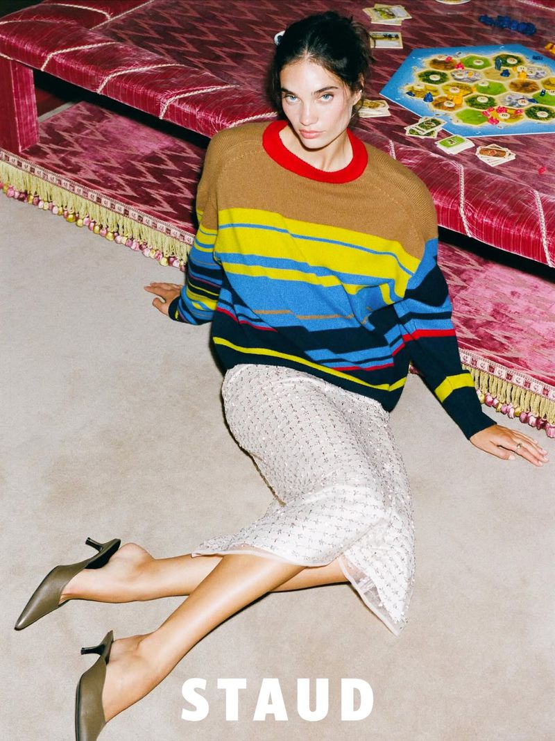 A woman poses on a patterned carpet wearing a striped sweater and beaded skirt, with a burgundy sofa and game in the background.