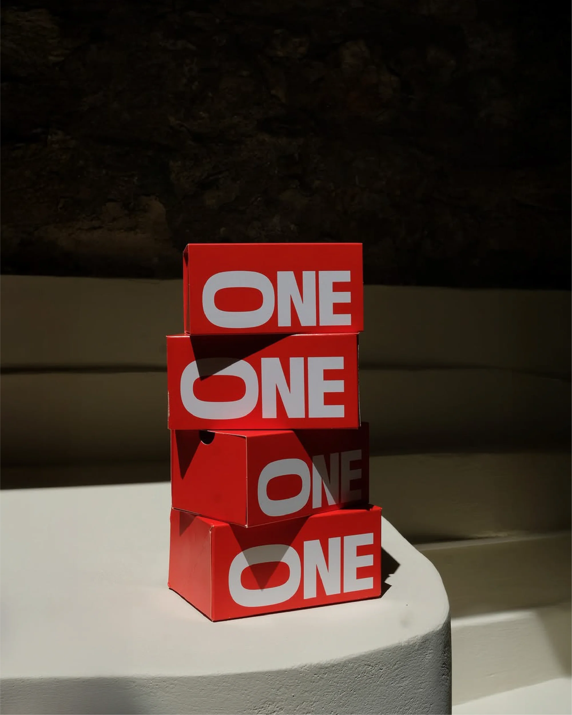 A stack of four red rectangular boxes, each labeled with 'ONE' in white, on a white surface against a dark wall, creating a minimalistic composition.