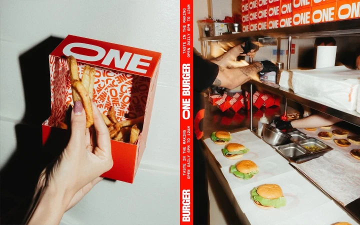 French fries are held over a red and white fast food container in a studio setting. Hamburgers are being prepared in the background.
