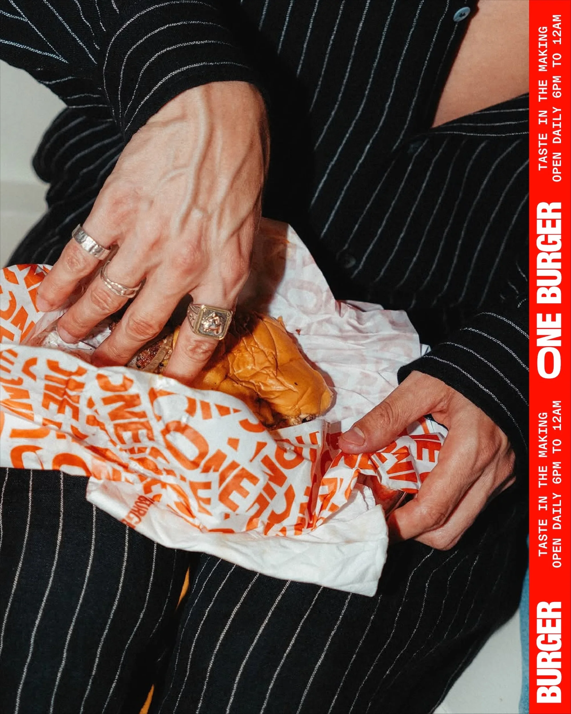 Close-up of a person in a striped shirt holding a burger wrapped in branded paper, featuring visible hand jewelry and saturated colors.
