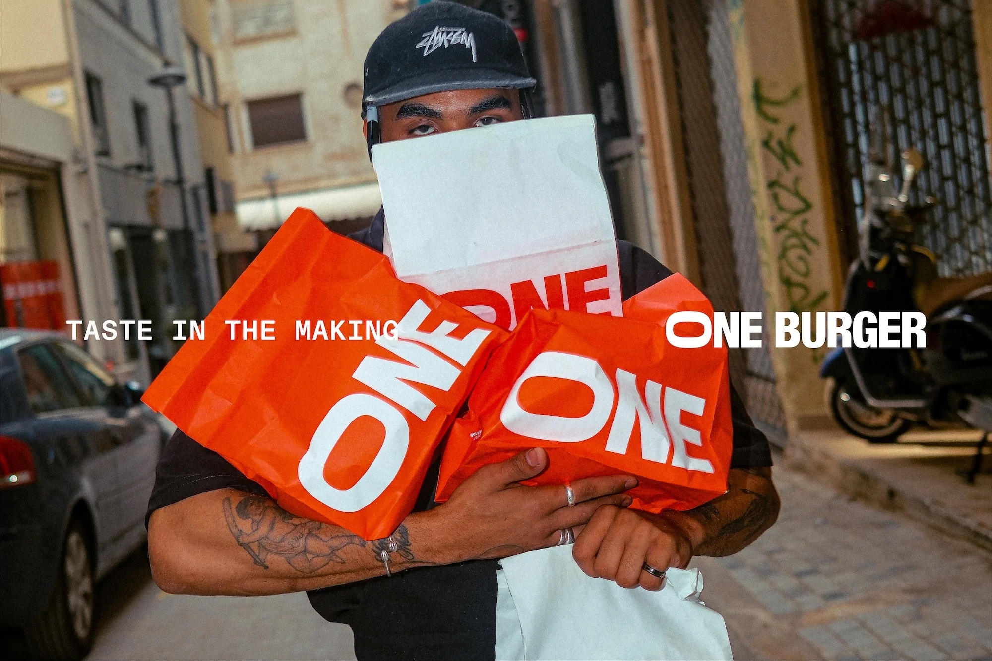 Adult male outdoors in an urban setting holding multiple shopping bags with 'ONE BURGER' text, wearing a black baseball cap and casual attire.