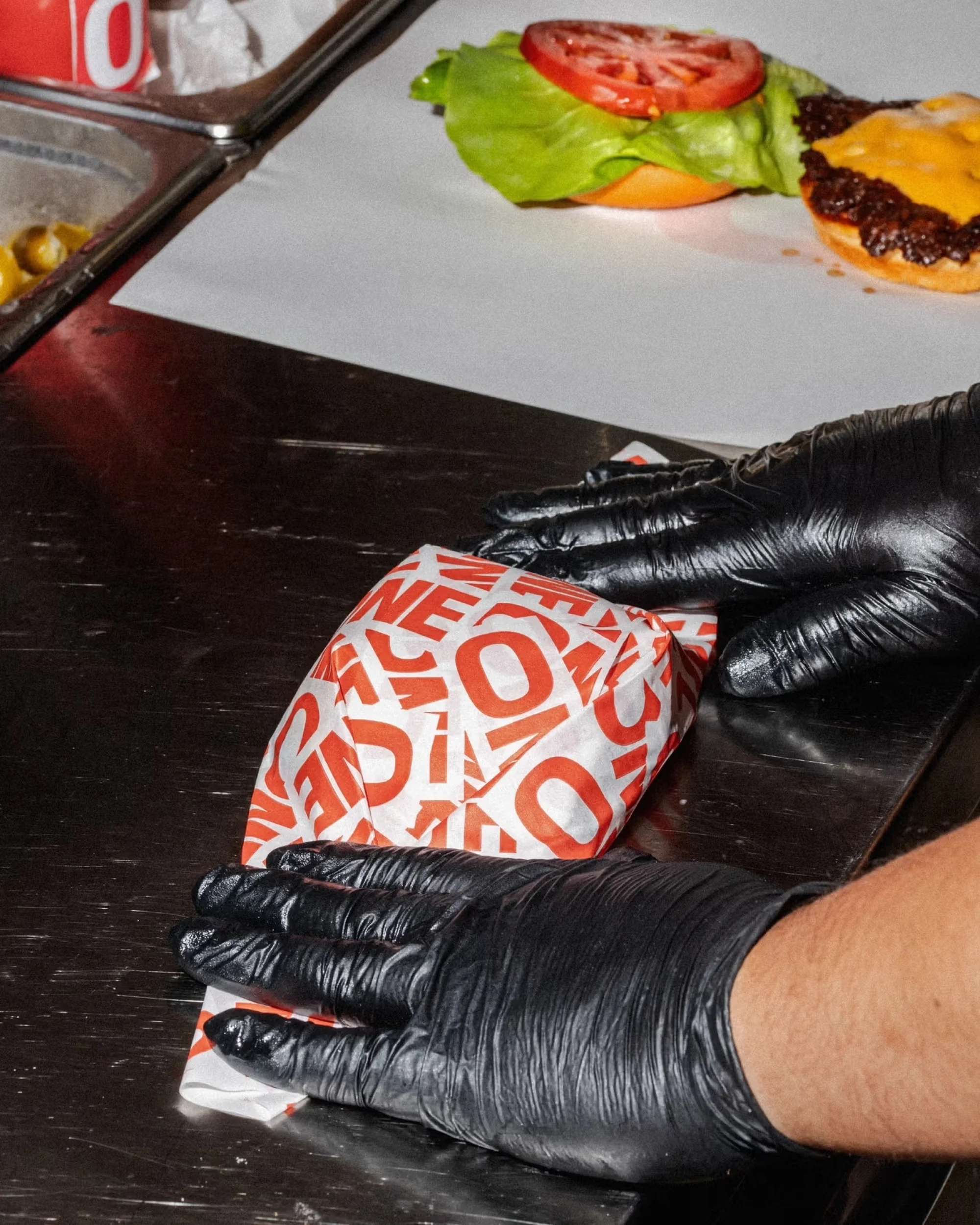 Burger preparation featuring a packaged burger being handled by a person wearing black gloves, with burger ingredients nearby.