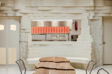 Modern restaurant interior featuring layered cream walls, a wooden table, and a service counter with red packaging.