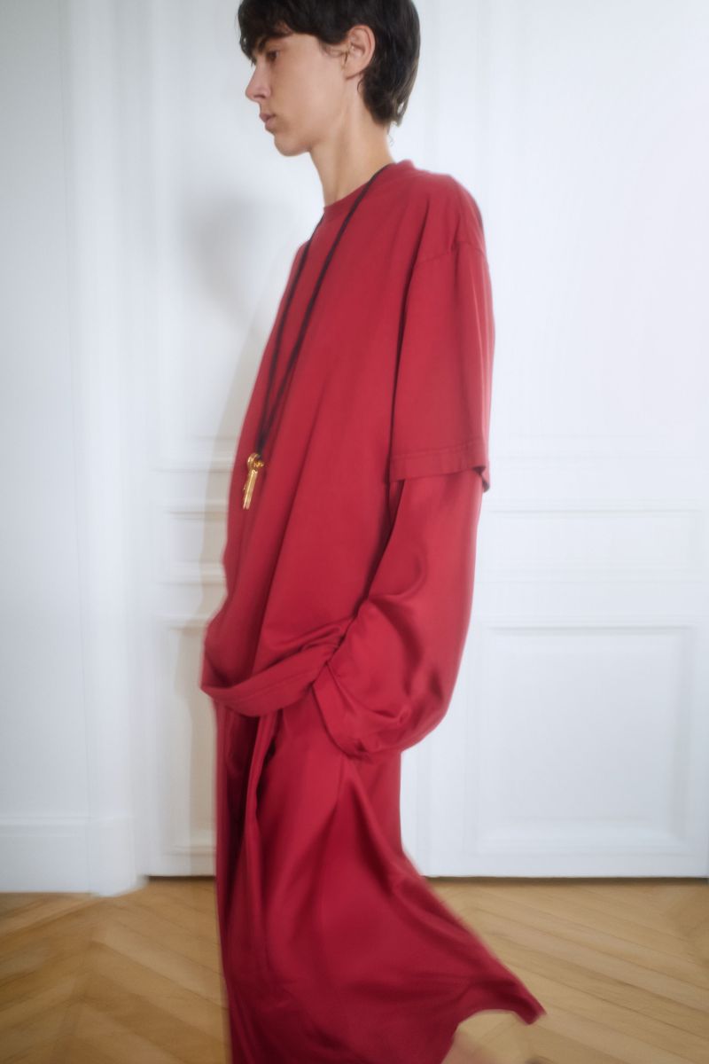 Profile shot of a person wearing an oversized, long-sleeved red shirt with matching long skirt, accessorized with a pendant necklace.