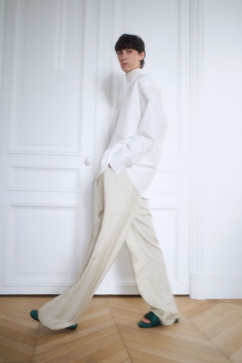 A slender person is walking in a brightly lit room, wearing a white button-down shirt and light pleated pants with green slide sandals.