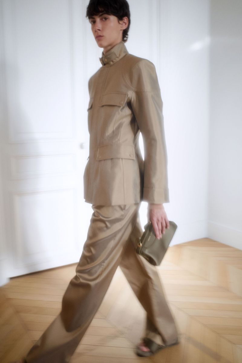 A young adult male is walking in a light beige two-piece suit and sandals, carrying a clutch, in a bright interior with wood floor.