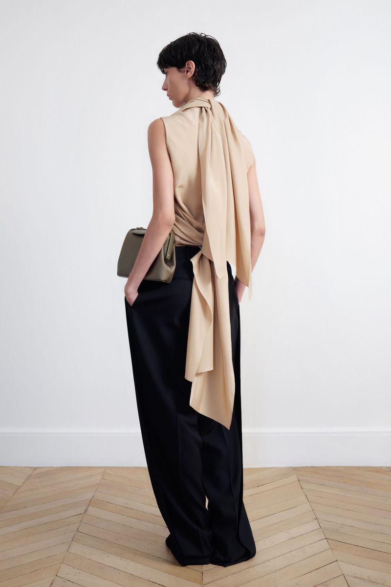 Back view of a woman in a tan top and black pants with a shoulder bag in a white interior with light wood floors.