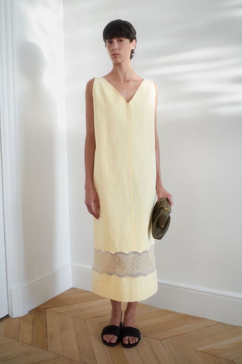 A person wearing a yellow dress and holding a clutch bag poses in an indoor setting with a wooden floor and white walls.