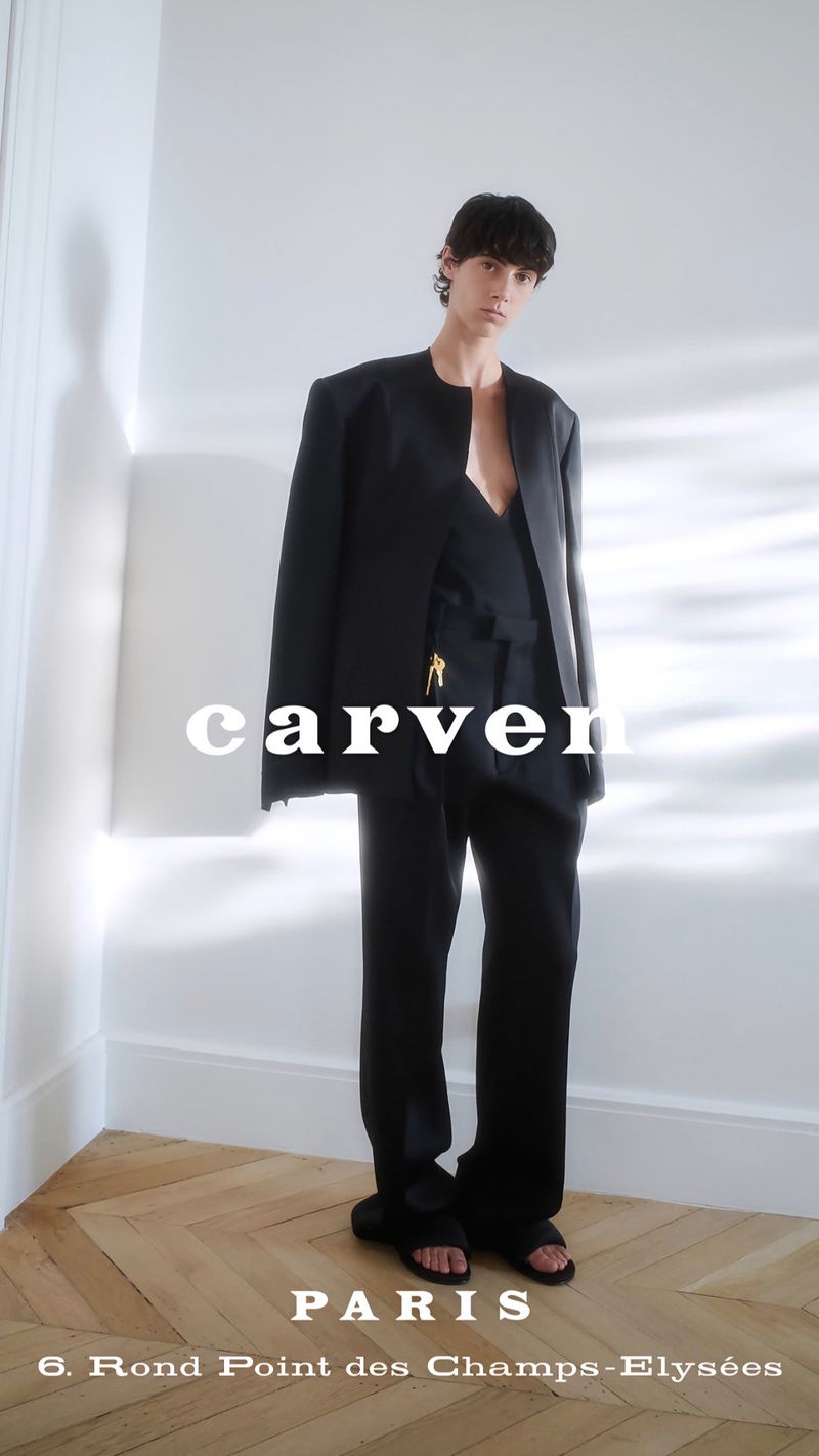 Model wearing a black jacket and trousers standing in a minimalist room with herringbone wooden flooring and white walls.
