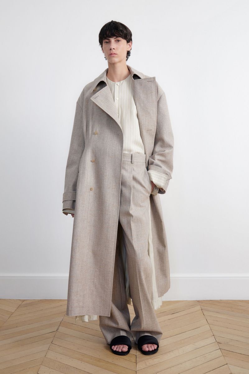 Fashion shot of a young adult posing indoors with a long trench coat, light colored trousers, and black sandals against a white wall.
