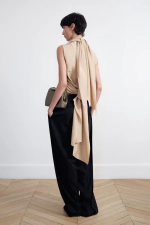 Model standing facing away wearing a draped beige top and black trousers holding a handbag