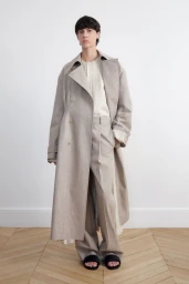 Model standing against a plain white wall wearing a beige trench coat and matching trousers over a cream shirt.
