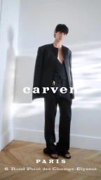 Model wearing a black jacket and trousers standing in a minimalist room with herringbone wooden flooring and white walls.