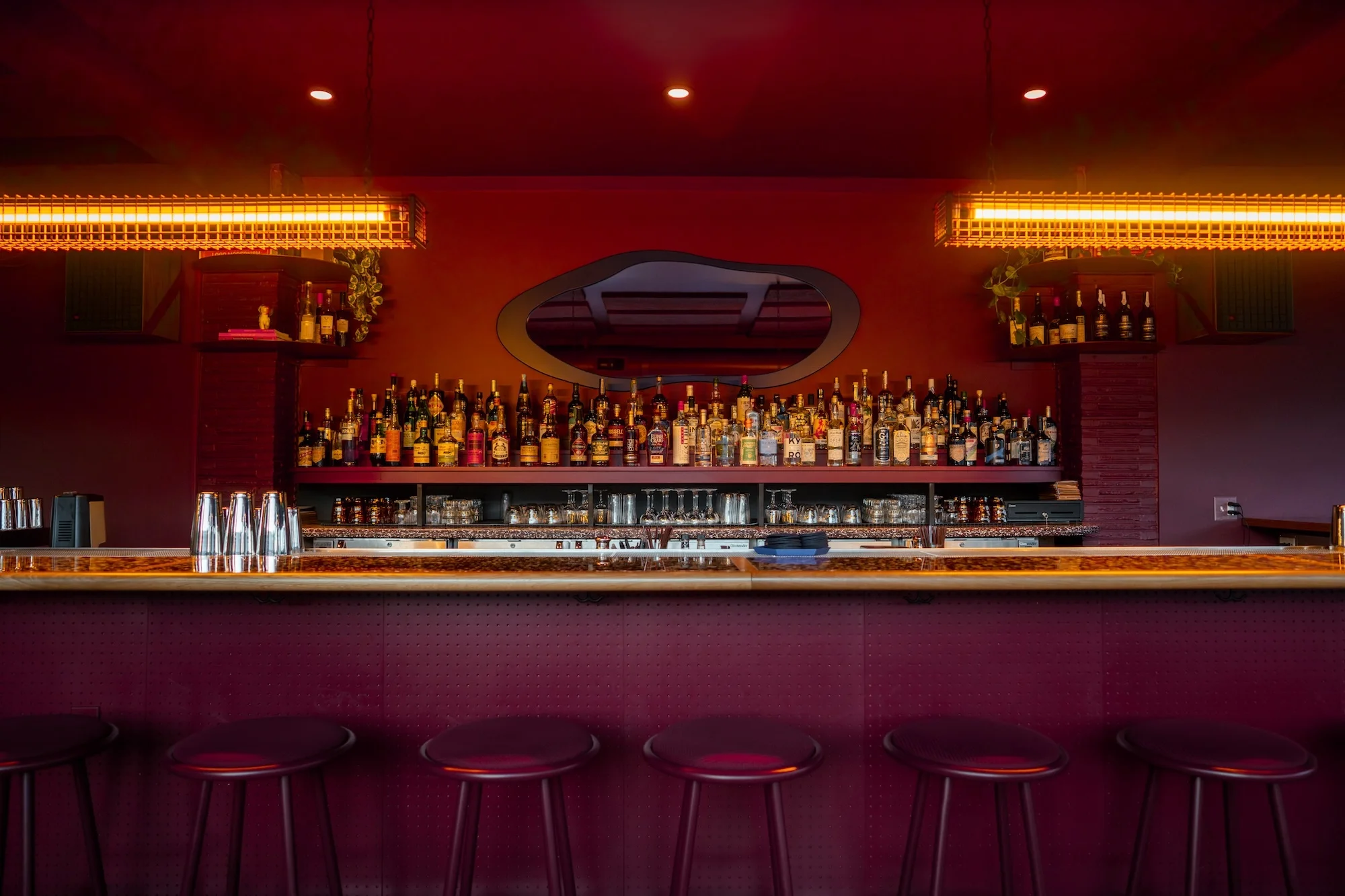 Interior shot of a bar featuring an array of liquor bottles behind the counter and stools lined up in front, lit by overhead lamps.