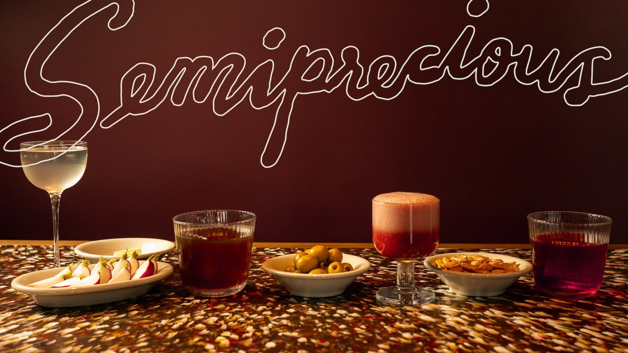 Several cocktails, bowls of snacks, and a cursive text graphic fill a tabletop composition under warm lighting against a brown backdrop.