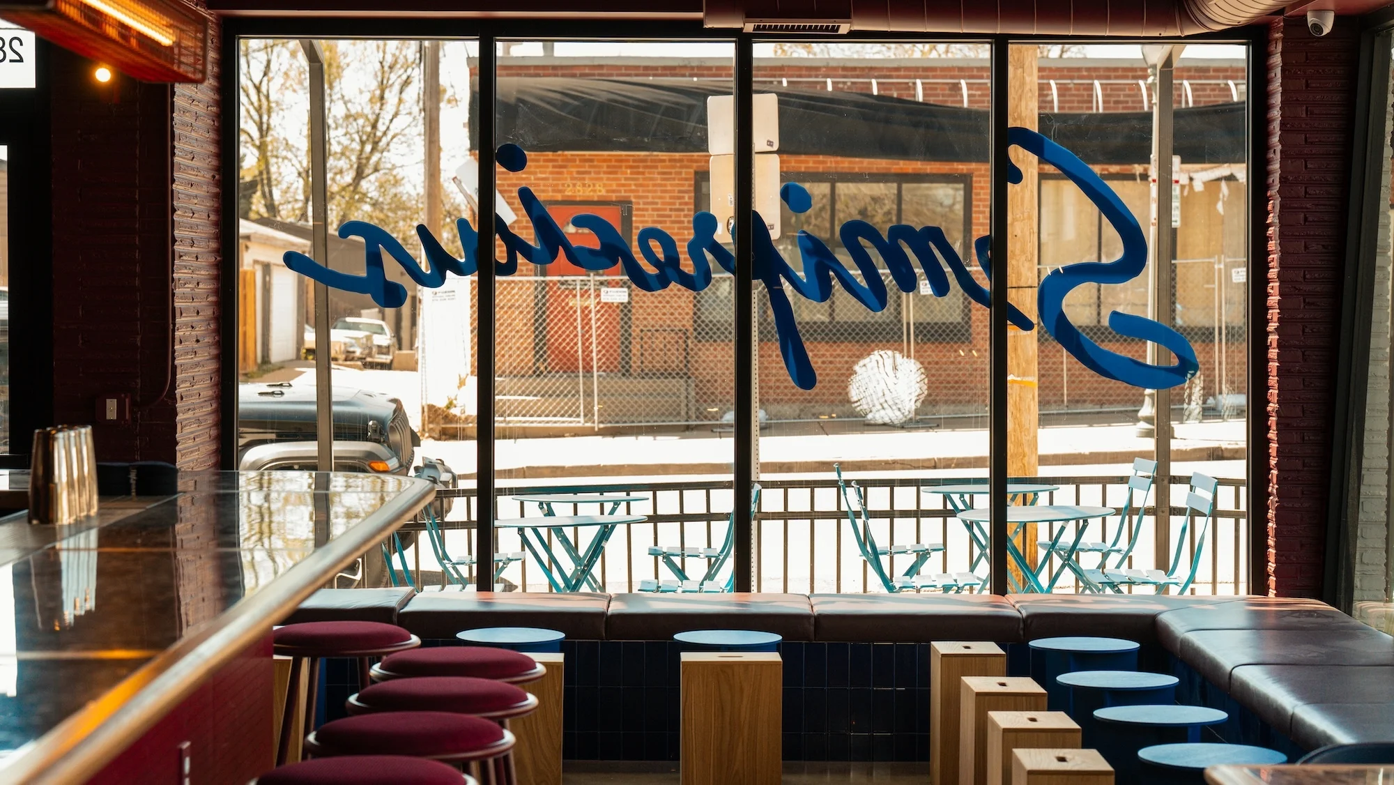 Interior view of a bar with stools, a bar counter, and a large window displaying outdoor seating and a brick building.