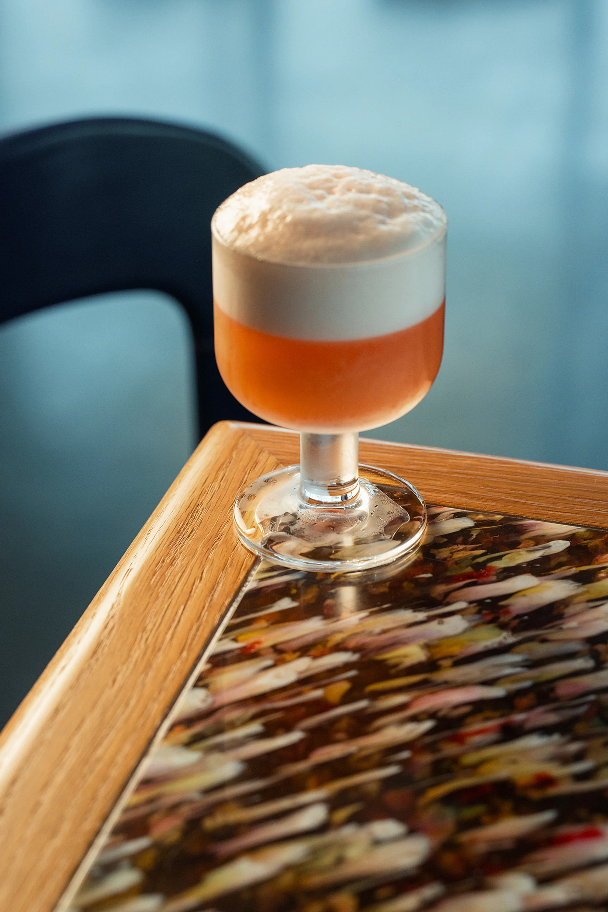 Refreshing peach-colored cocktail topped with foamy white head sits on a patterned table near a dark chair and bright window.