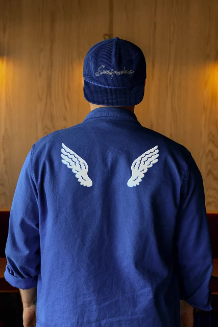 Back view of a person in blue shirt and cap with white wing designs, standing in front of a wooden wall; fashion and textile design.