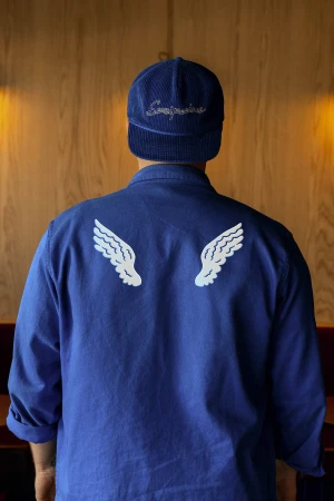 Back view of a person in blue shirt and cap with white wing designs, standing in front of a wooden wall; fashion and textile design.