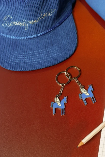 Two winged horse keychains sit on a red surface with a blue cap and a pencil and paper providing a colorful, detailed product shot.