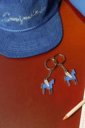 Two winged horse keychains sit on a red surface with a blue cap and a pencil and paper providing a colorful, detailed product shot.