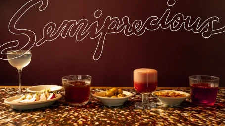 Cocktail glasses, small plates of radishes, olives, and snacks arranged on a patterned tabletop against a dark wall with white lettering