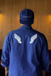 Rear view of a person wearing a blue corduroy cap and blue button-up shirt with white angel wing graphics on the back