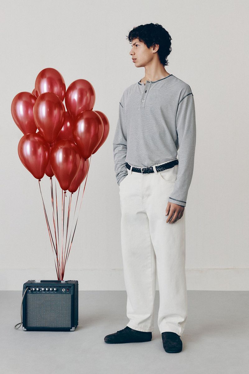 A young adult male stands indoors next to red balloons and a small amplifier. He wears a casual long-sleeved shirt and white pants.