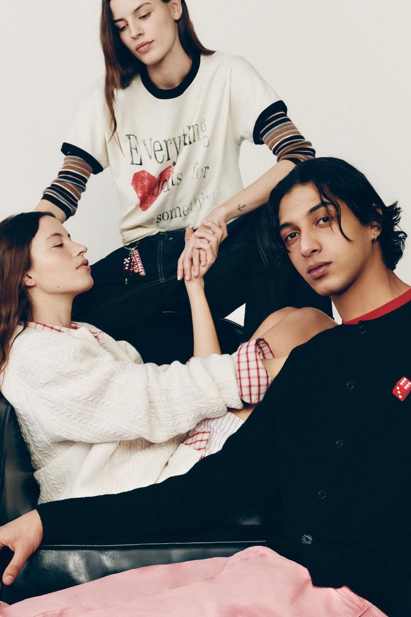Three fashion models pose indoors on a black sofa wearing modern clothing with a white background, fashion editorial style.