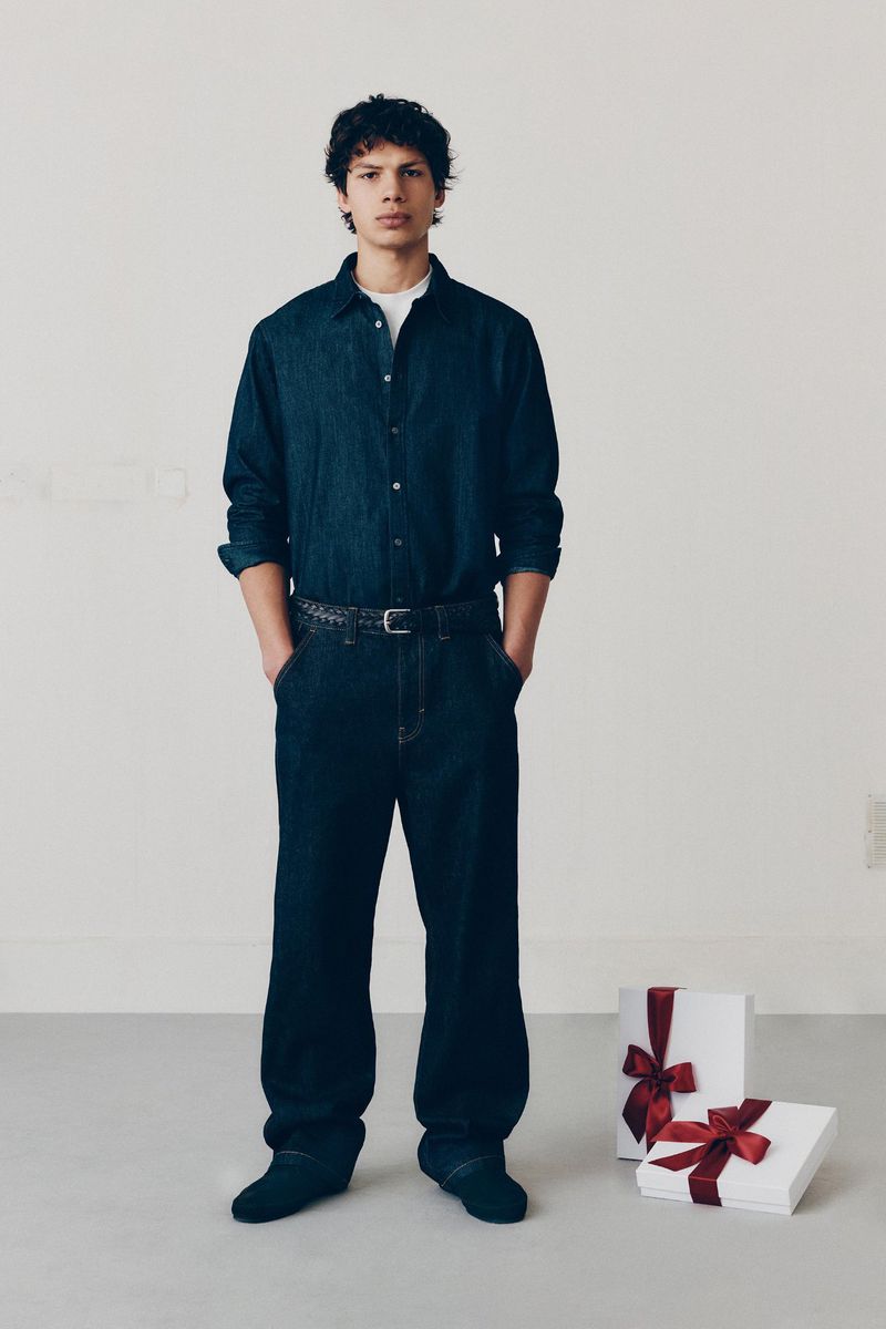 A young adult male in a blue denim outfit stands in front of a cream background with two gift boxes wrapped with red ribbon on the floor.