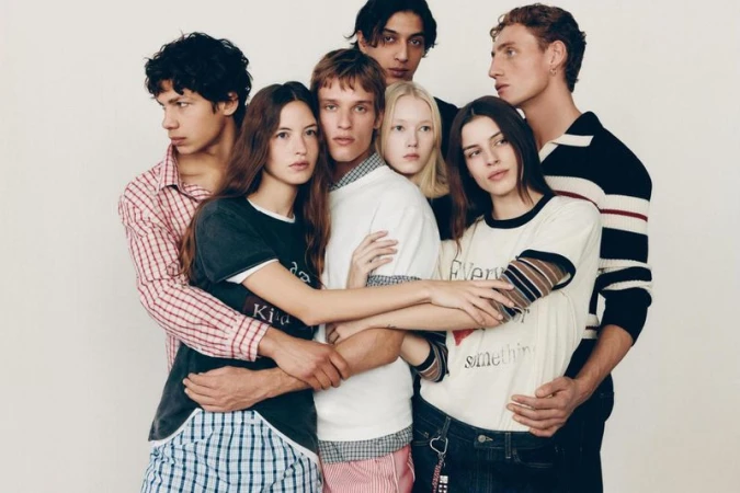 A group of six young adults huddle closely together in a fashion-style photo against a plain backdrop, wearing casual clothing.