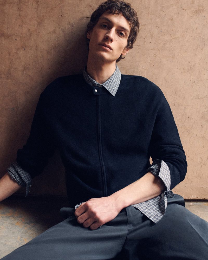 Studio shot of a young adult man in a navy zip-up sweater and gray trousers posing in front of a textured brown wall. 
