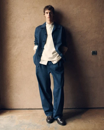 Man in relaxed pose, wearing blue shirt jacket and pants, cream shirt and brown shoes, against a tan textured wall backdrop.