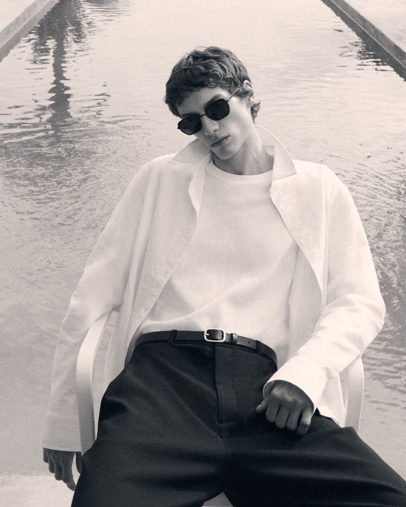 Monochrome shot of a male model in sunglasses, a white shirt, and dark pants, posing by the water outdoors with a shallow depth of field.