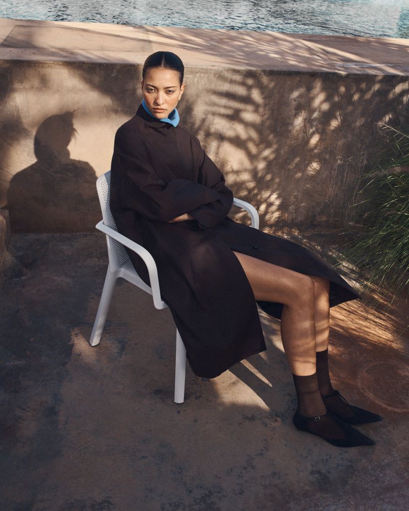Elegant woman in a long black dress sitting on a white chair near a swimming pool with sunlit shadows on a textured wall.