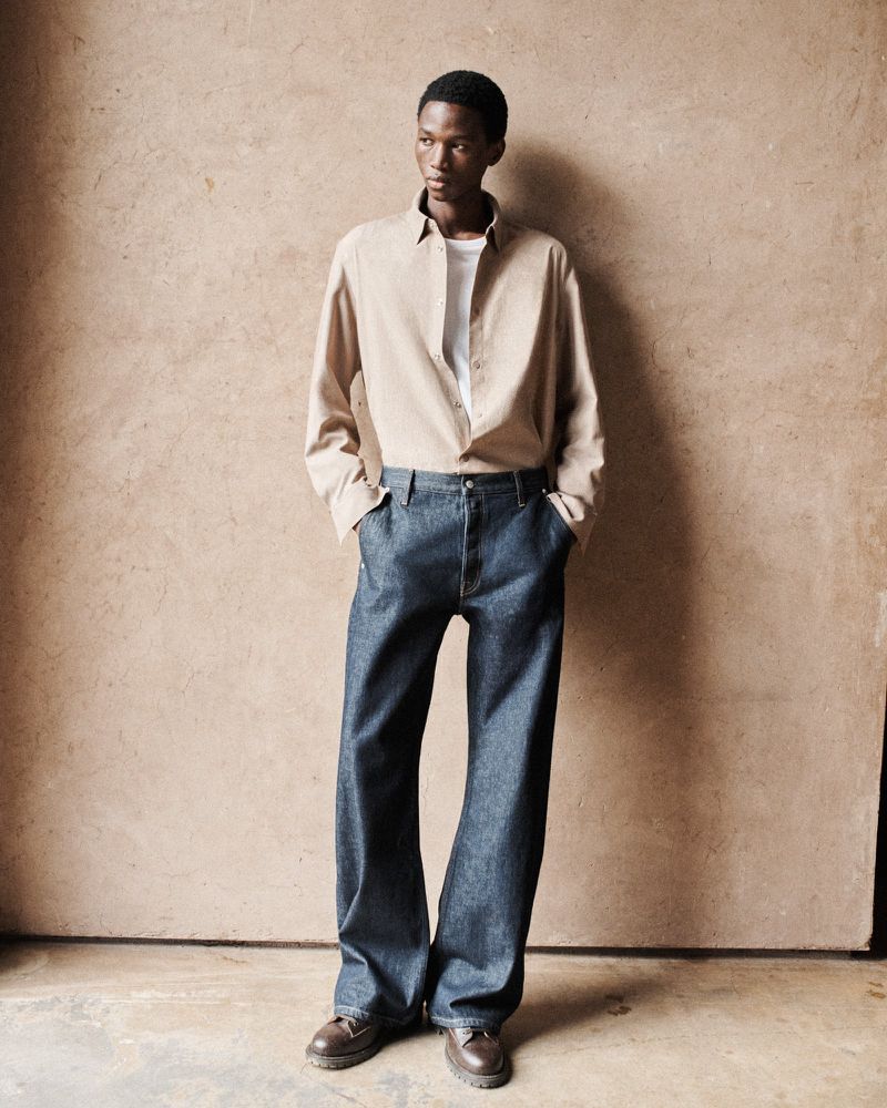 Full-length shot of a slender, dark-skinned male model in a tan collared shirt and flared dark-wash denim jeans, posing against a textured beige wall.
