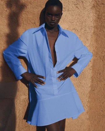 A Black female model poses in a blue, oversized, collared, long-sleeve dress with a flounce, against a brown textured wall with soft shadows.