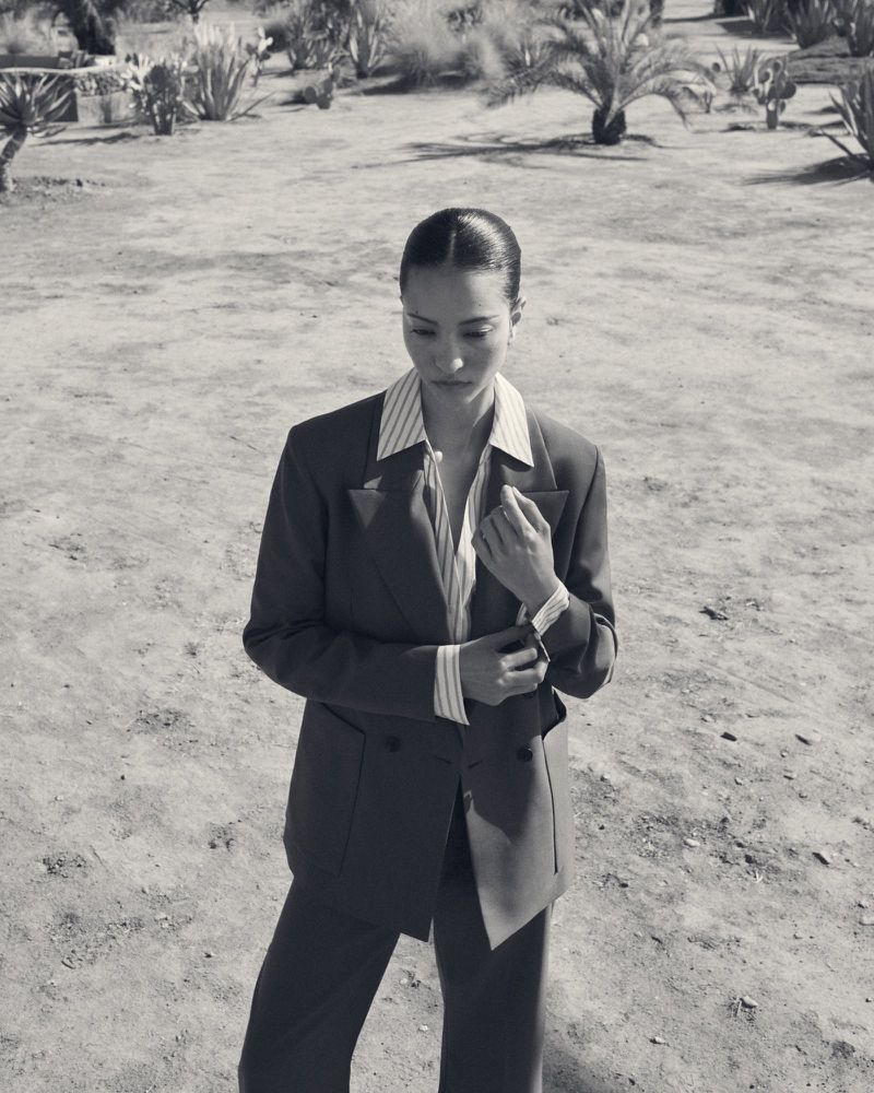 Monochrome portrait of a young adult woman in a suit, buttoning her sleeve in an arid outdoor setting with sparse vegetation.