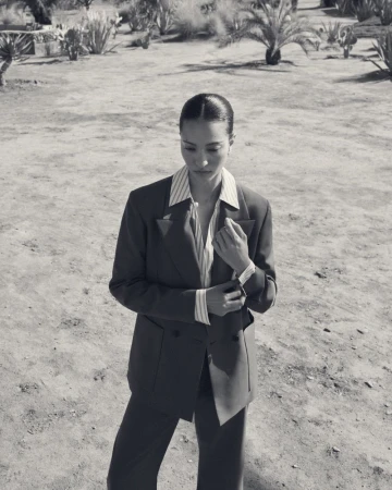 Monochrome portrait of a young adult woman in a suit, buttoning her sleeve in an arid outdoor setting with sparse vegetation.