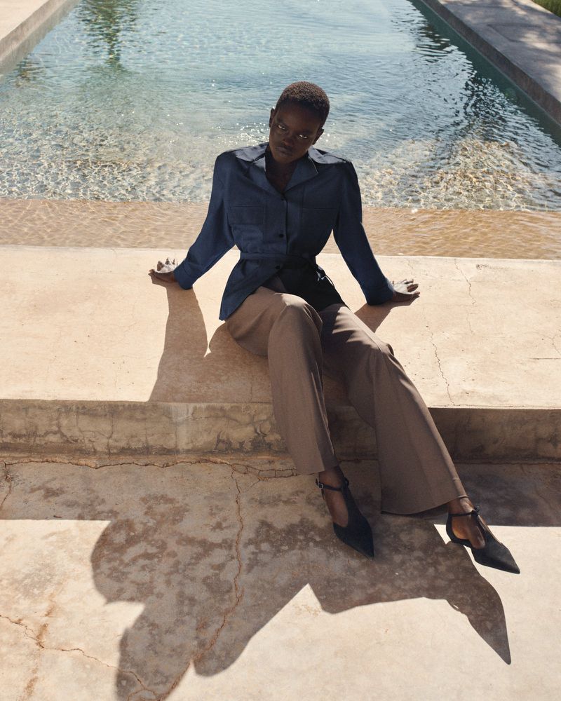 Fashion shot of an African woman in a blue top and beige pants, sitting by a pool on a sunny day, creating shadows and soft light.