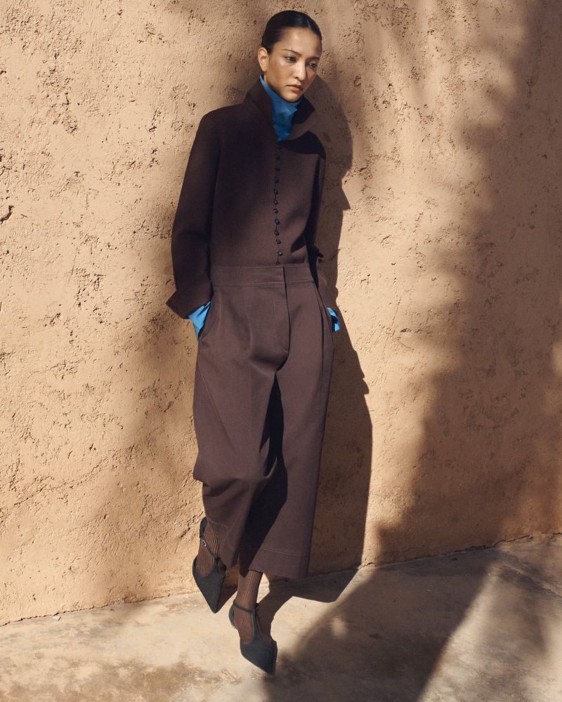 A fashion shot features a woman in a brown jumpsuit and black heels posing against a textured wall with dappled light and shadows.