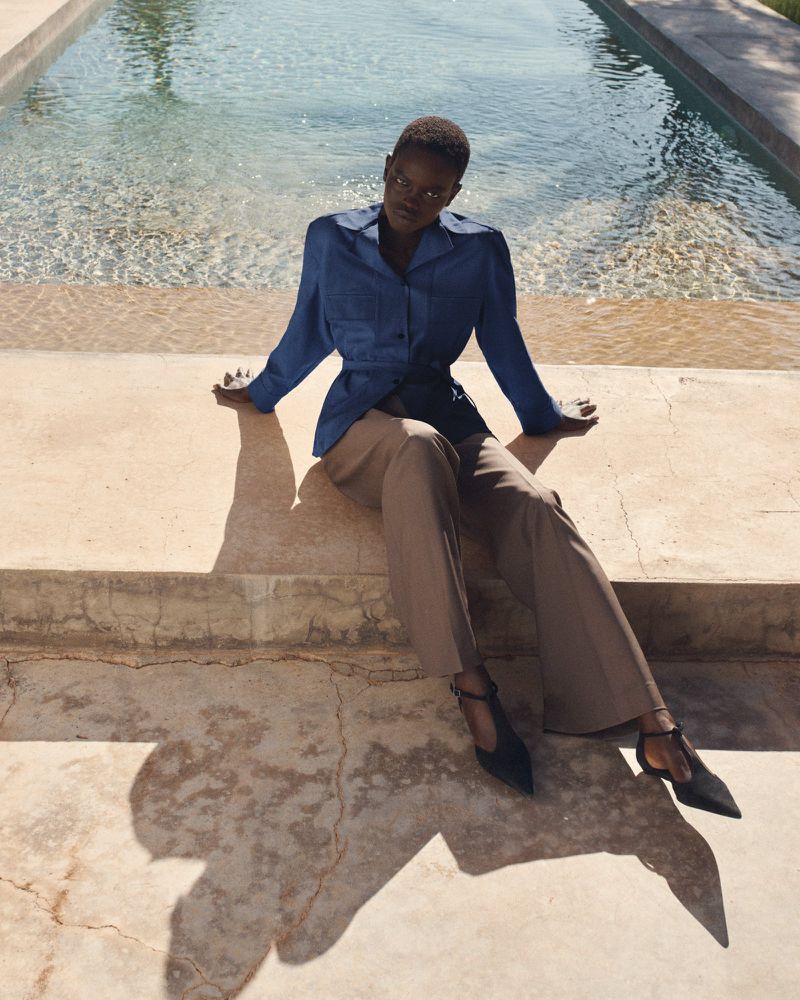 A dark-skinned woman poses in a blue button-down shirt and wide brown pants, seated on concrete steps near a pool, casting a shadow.