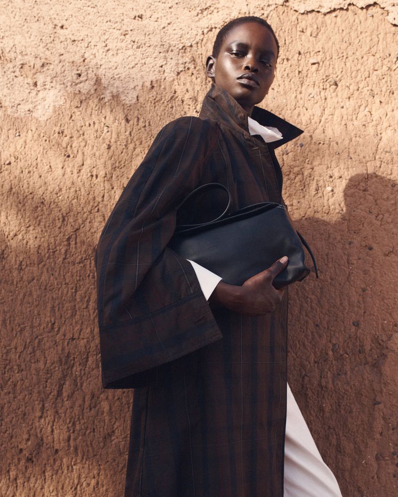Fashionable woman in a coat holds a black purse in an outdoor setting with a textured wall backdrop, elegant pose, and soft lighting.