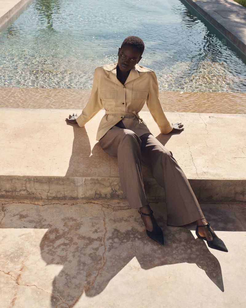 Model wearing a tan jacket and trousers sitting by an outdoor swimming pool in bright sunlight for COS