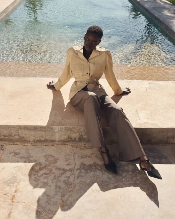 Model wearing a tan jacket and trousers sitting by an outdoor swimming pool in bright sunlight for COS