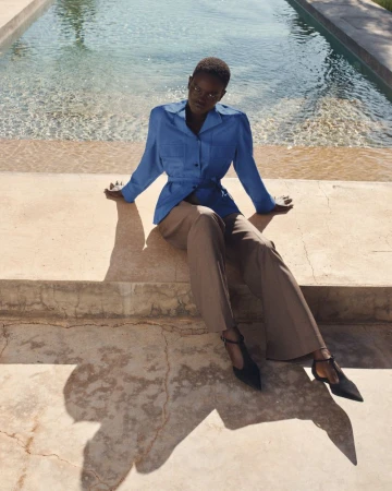 Model seated poolside wearing a blue collared shirt and wide trousers for the COS Pre-Spring 2026 lookbook by Pablo Di Prima.